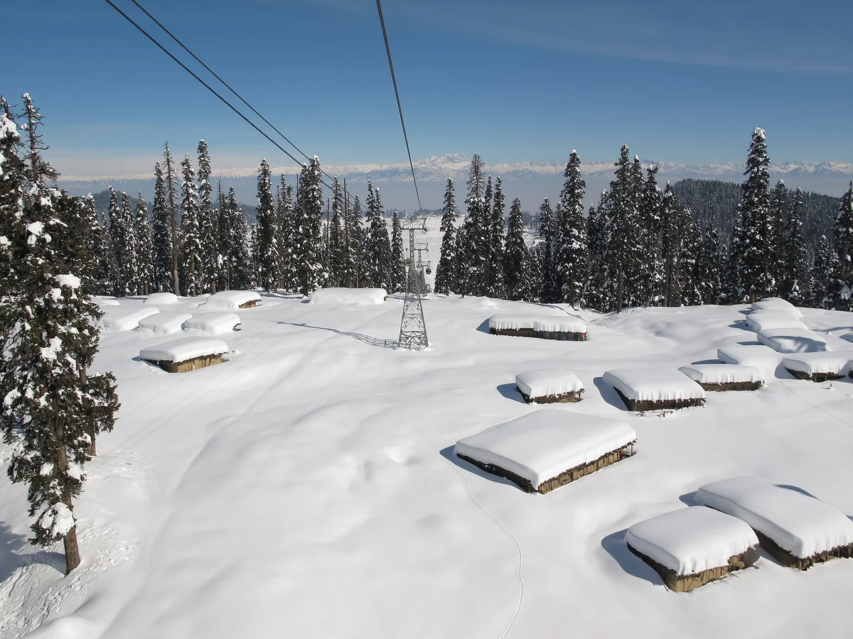 Snow-covered huts and pine trees below the Gulmarg Gondola cable, seen from a gondola cabin