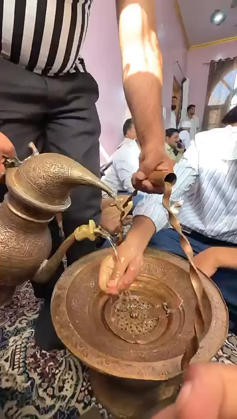 A diner rinsing his hands over an engraved tash-t-nari basin as water is poured from the jug, with guests seated on the floor behind at a Kashmiri wedding