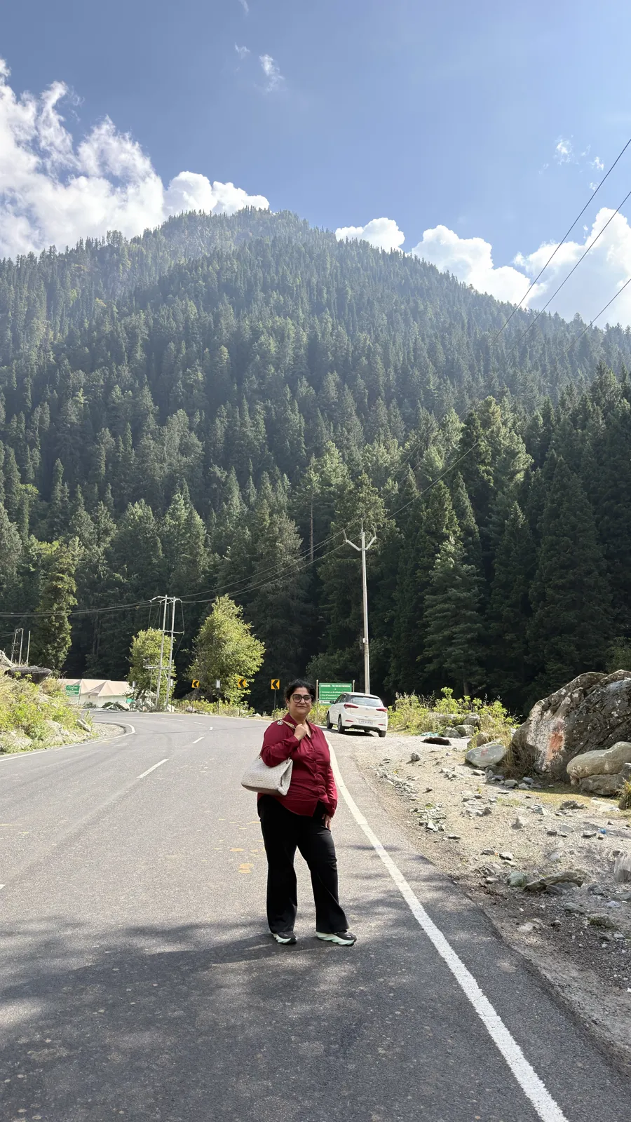 A woman from our ladies group in a red top standing on a mountain road with a pine-covered hill and blue sky behind her