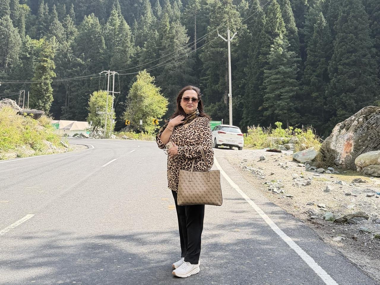 A woman in a leopard-print top and sunglasses standing on a smooth tarred mountain road, with thick pine forest on the hillside behind, on the way to Sonmarg