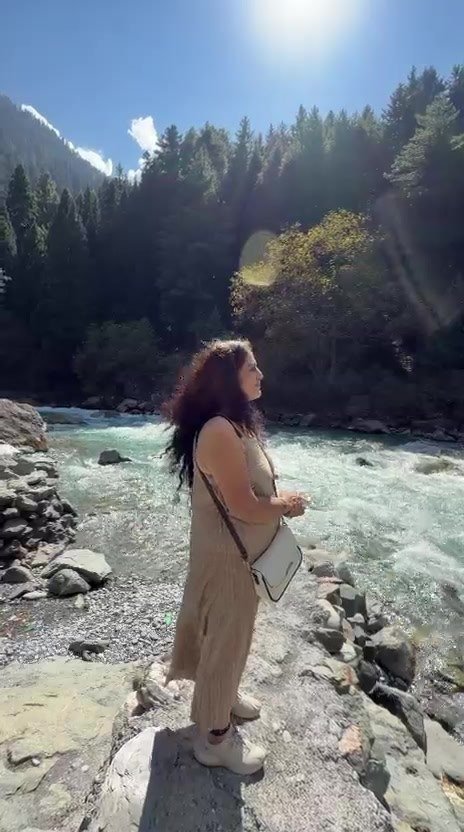 A woman standing on the rocky edge of a rushing river, with pine forest, a yellowing autumn tree and mountains behind under a sunny sky