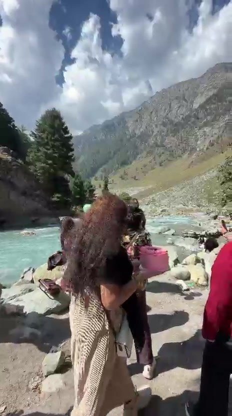 A group of women with a pink cake on a riverbank, with pine trees, a steep mountainside and big white clouds behind