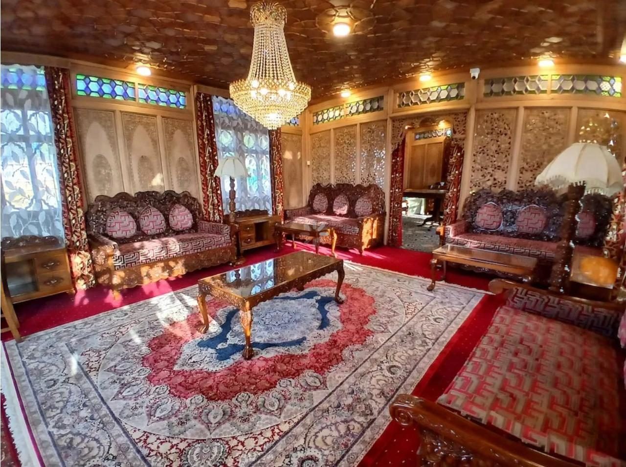 Carved wooden sitting room on a Srinagar houseboat with a crystal chandelier, carved sofas with red patterned cushions, a large patterned carpet and stained-glass window panels