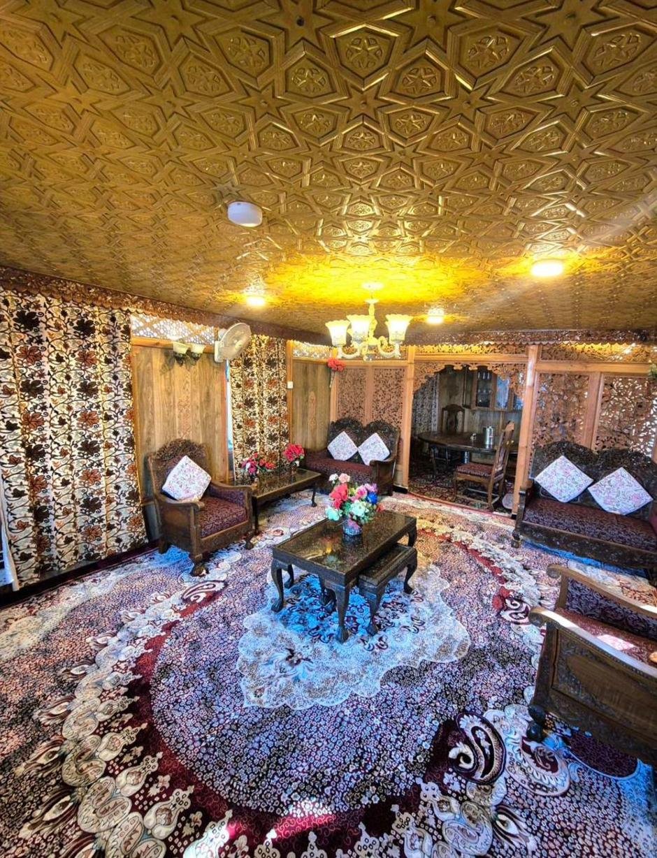 Houseboat sitting room with a star-pattern carved wooden khatamband ceiling, carved armchairs and sofas with white cushions, flowers on a low table and a large round carpet