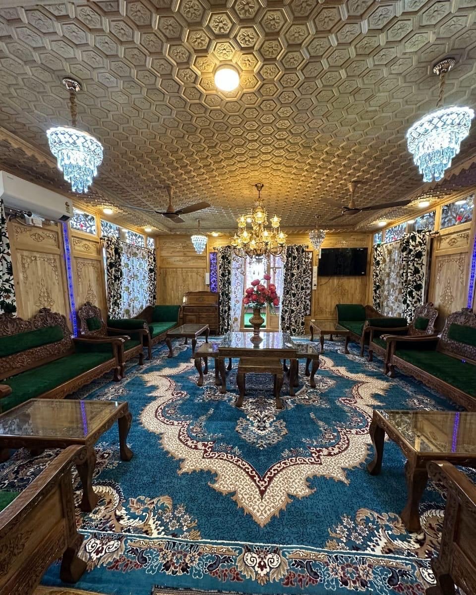 Long houseboat lounge with a carved wooden ceiling, crystal chandeliers, green velvet sofas along both walls, carved glass-topped tables and a blue patterned carpet