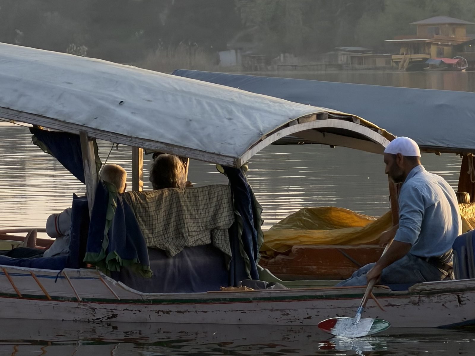 A boatman in a white cap paddling a covered shikara with guests on calm water at sunrise, with trees and a wooden building on the far shore