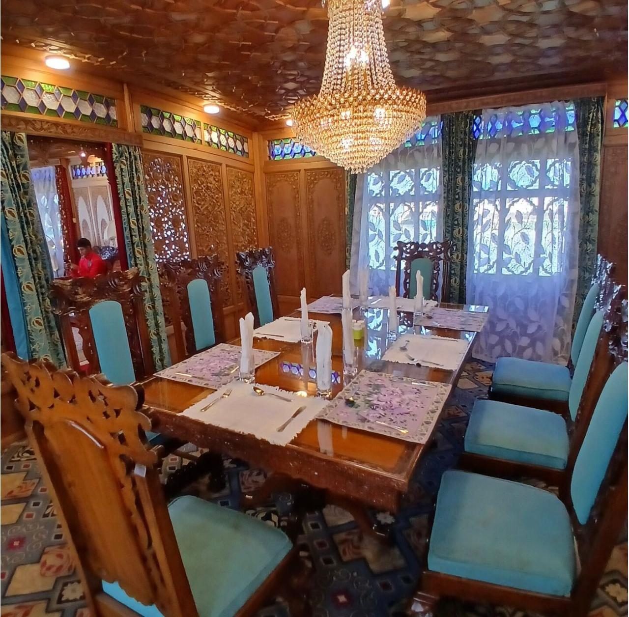 Houseboat dining room with a long carved wooden table laid with place mats and napkins, turquoise upholstered carved chairs, a crystal chandelier and lace curtains