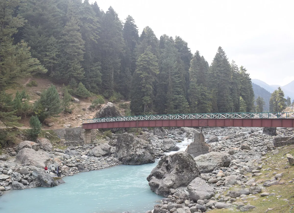 The Lidder river flowing over grey rocks through the Pahalgam valley, with a red footbridge and pine-covered slopes