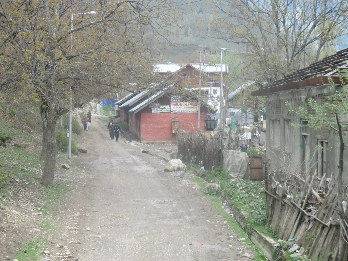 A dirt lane running between wooden and brick village houses in Pahalgam, with bare trees and hills behind