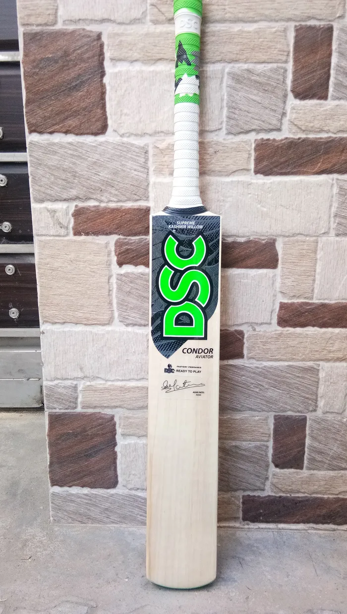 A new Kashmir willow cricket bat with a green and white handle standing against a stone wall