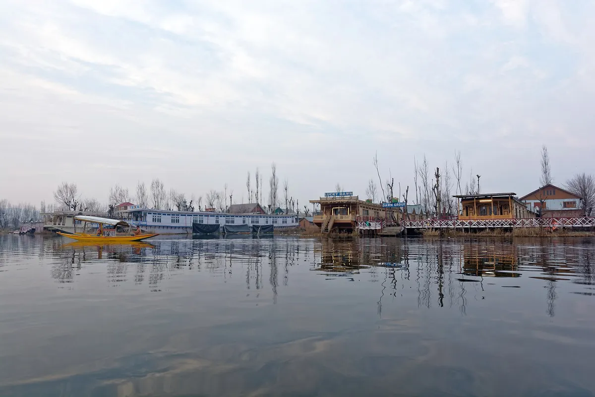 Wooden houseboats along the shore of Dal Lake in Srinagar on a grey winter day, with a yellow shikara on the water and bare trees behind