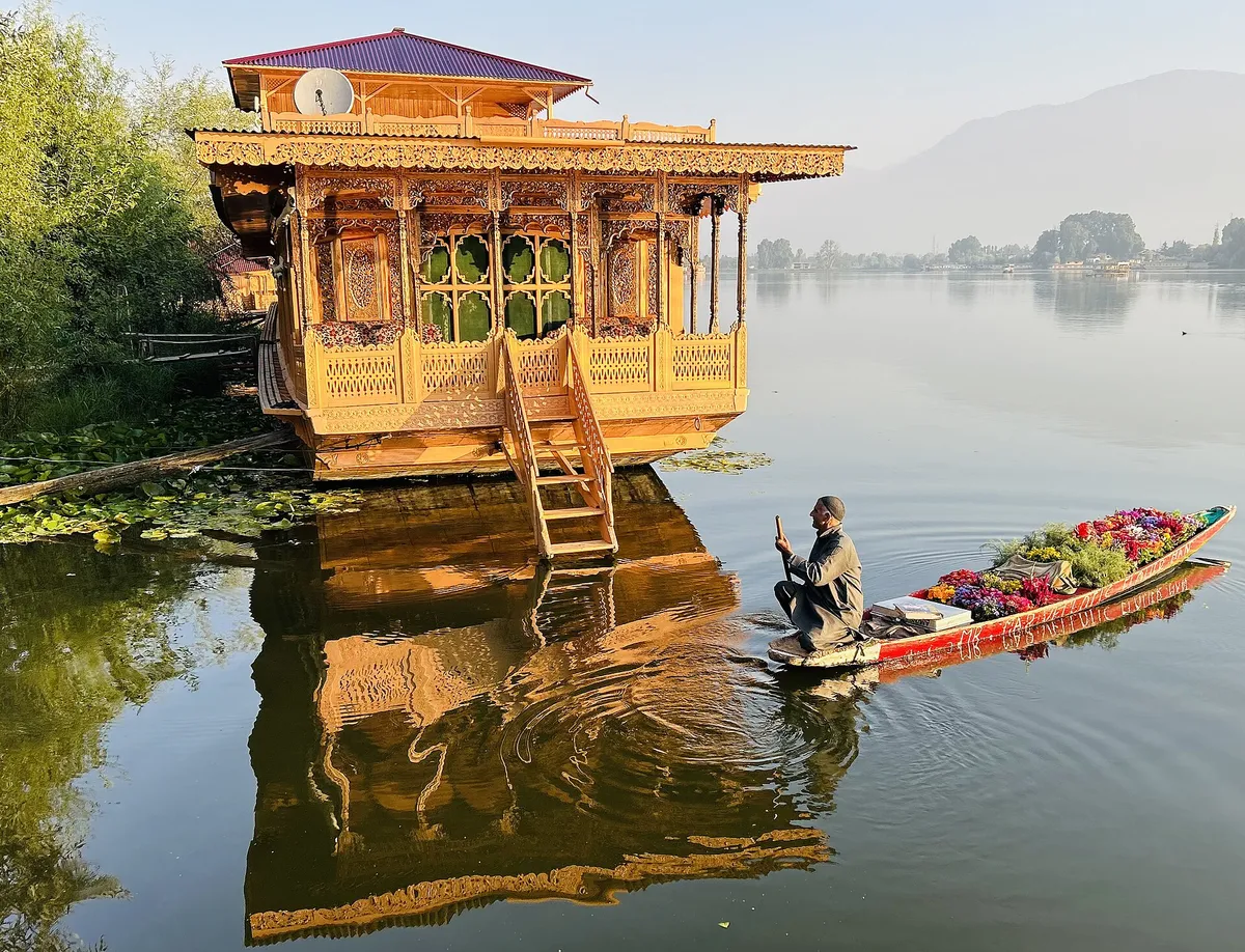 A carved wooden houseboat on Nigeen Lake in Srinagar with a flower seller's shikara alongside and its reflection in still water