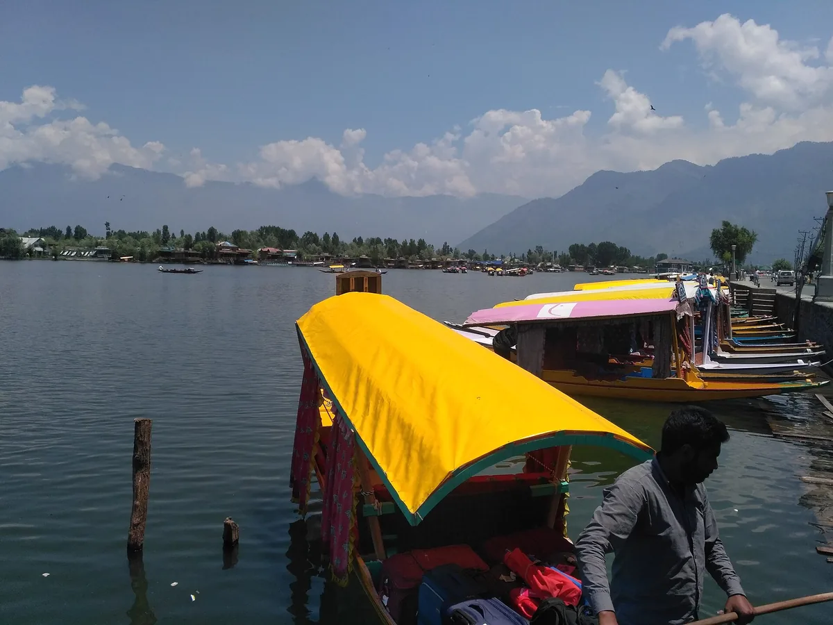 Colourful shikaras docked beside Boulevard Road on Dal Lake in Srinagar, with luggage on a boat and mountains across the water