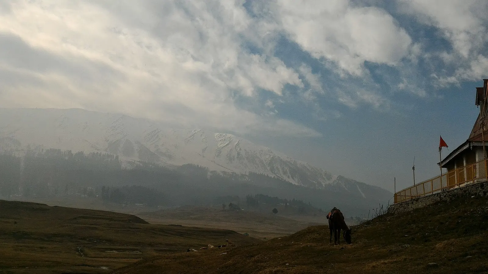 Snow on the mountains above Gulmarg in late November, with the meadow below still brown and a horse grazing near a wooden hut