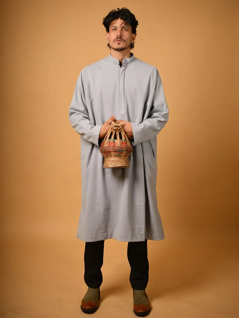 A young Kashmiri man in a grey woollen pheran holding a wicker kangri fire pot