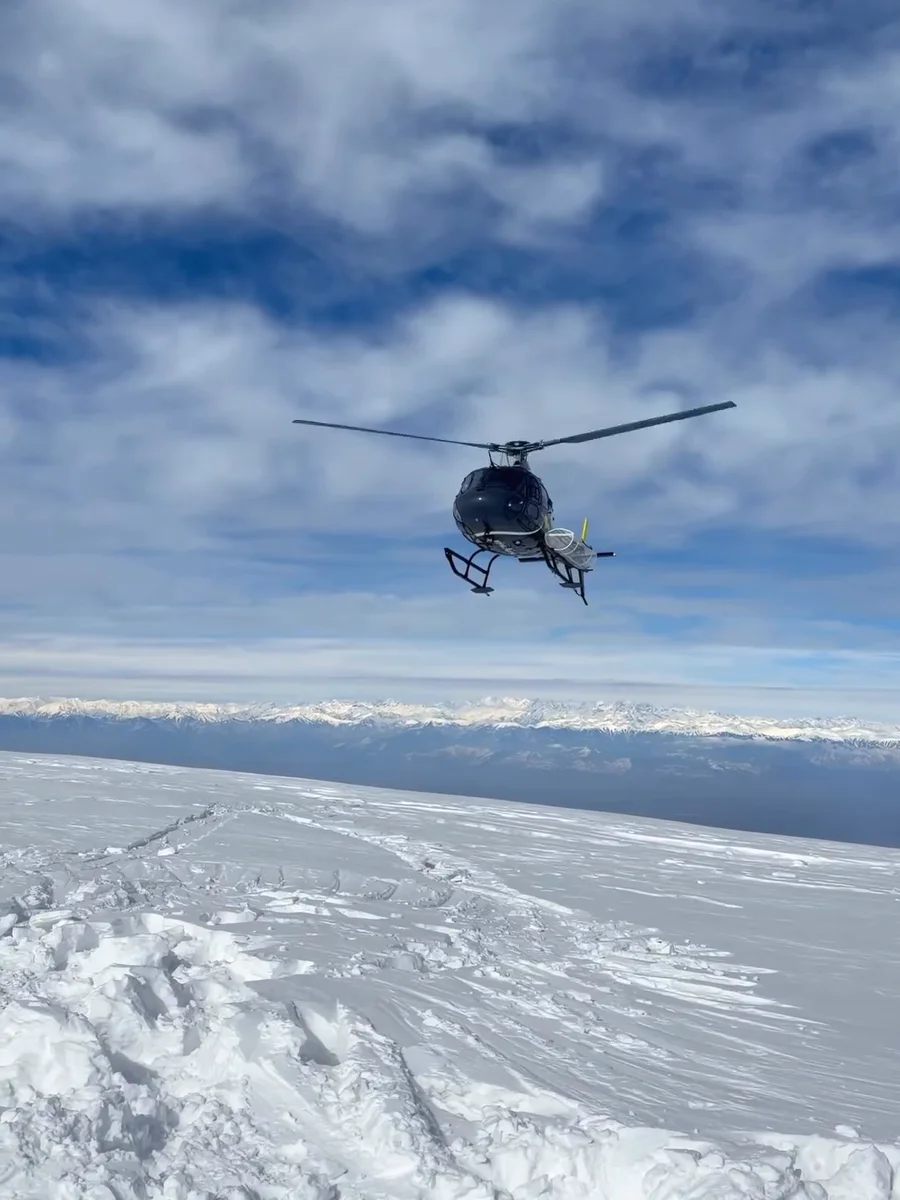 A helicopter flying in towards a snowfield above Gulmarg under a blue sky with white clouds, from our reel