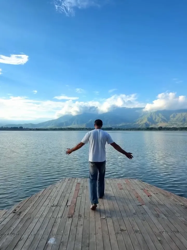 Zahid walking with arms open along a wooden jetty over Dal Lake under a blue sky with clouds over the mountains