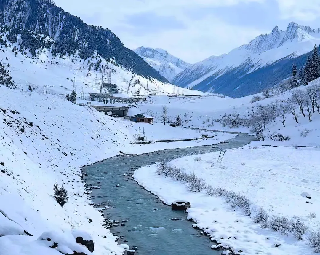 A green river winding through a snow-covered Kashmir valley with white peaks and blue sky