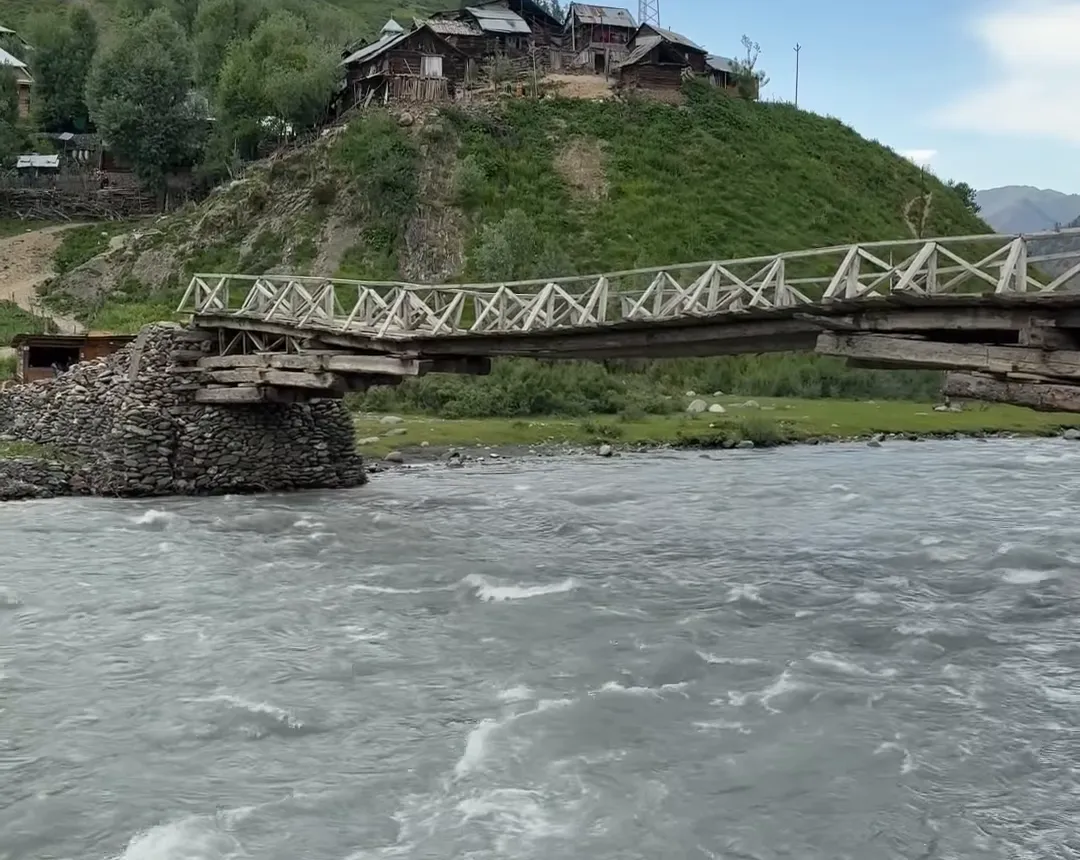 A footbridge over the fast Kishanganga river below wooden houses of Bagtor village in Gurez