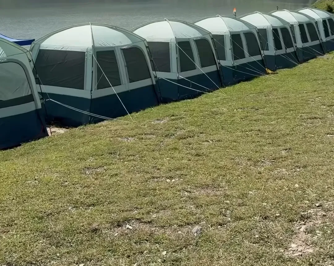 A row of green dome tents on a grassy riverbank in Gurez with forested hills behind