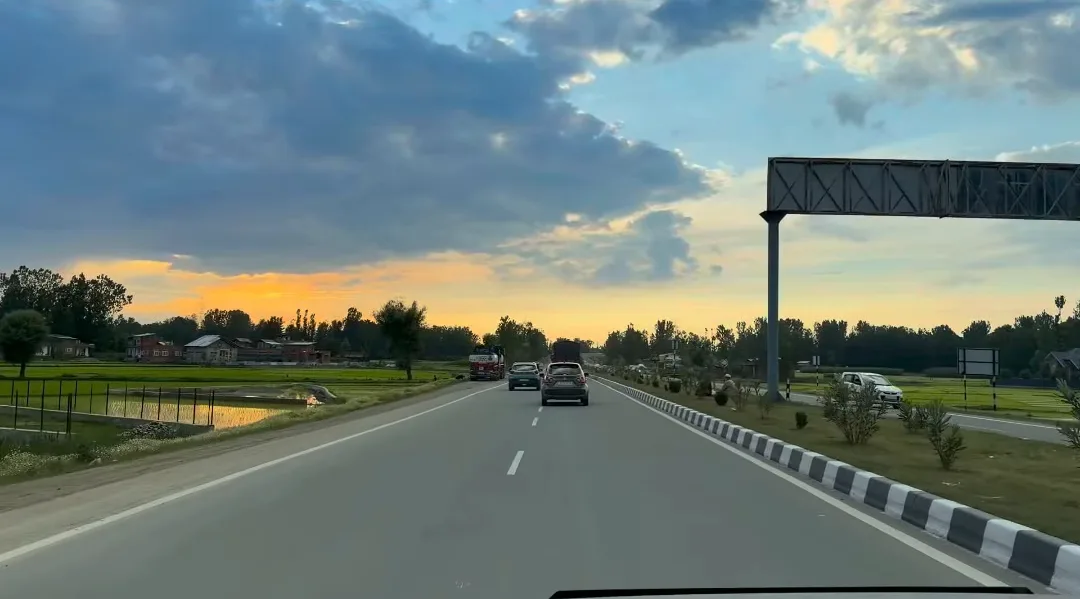 A four-lane highway in the Kashmir valley at sunset with green paddy fields and an overhead sign gantry