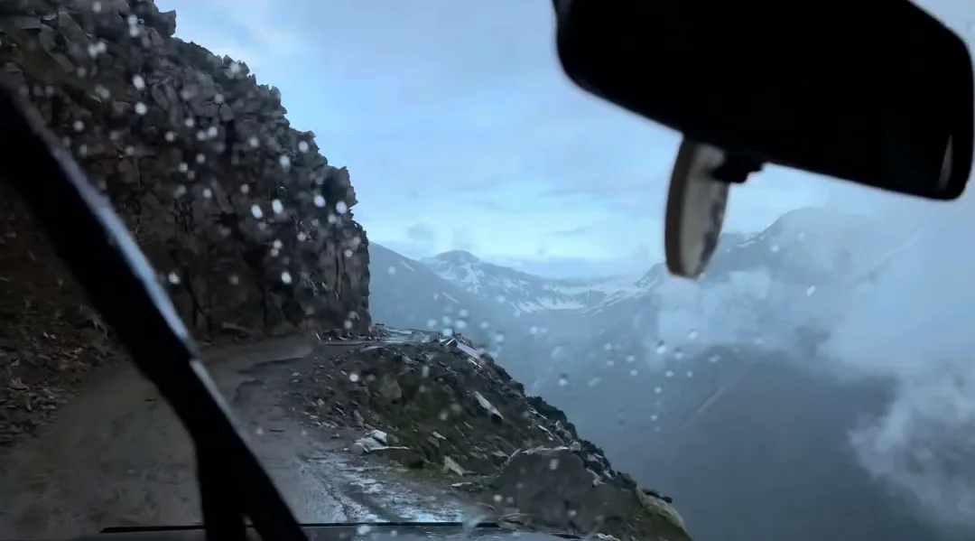 View through a car windscreen of a narrow mountain road with a rock wall on one side and clouds over the peaks
