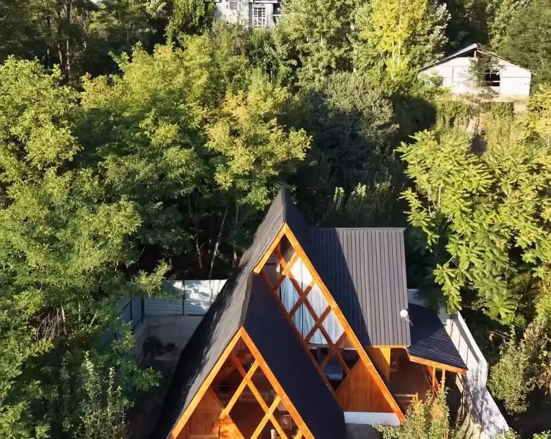 Aerial view of a wooden A-frame cottage with a glass front among green trees in Kashmir