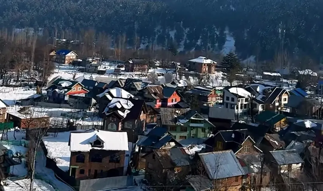 Snow-covered rooftops of a Kashmir town with pine hills behind, from our winter reel